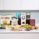 Gourmet Delights Hamper. A selection of gourmet snacks is displayed on a light-colored countertop. Items include a bag of Keogh's cheese and onion crisps, various cheeses, crackers, grapes, chocolate bars, and cookies. Several product boxes are arranged in the background, showcasing brands like Forest Feast and Lily O'Brien’s. The overall presentation suggests a curated food assortment ideal for entertaining or snacking. Gourmet Delights Hamper. A selection of gourmet snacks is displayed on a light-colored countertop. Items include a bag of Keogh's cheese and onion crisps, various cheeses, crackers, grapes, chocolate bars, and cookies. Several product boxes are arranged in the background, showcasing brands like Forest Feast and Lily O'Brien’s. The overall presentation suggests a curated food assortment ideal for entertaining or snacking.